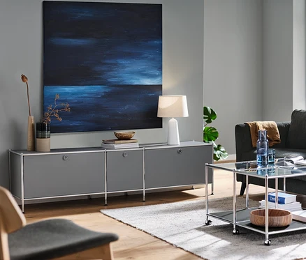 Šedá nízká skříňka CN3 se 3 výklopnými dvířky, stolní lampa s bílým textilním stínidlem a antracitová pohovka pro 3 osoby v obývacím pokoji. Také konferenční stolek CN3 s kolečky a lounge židle.