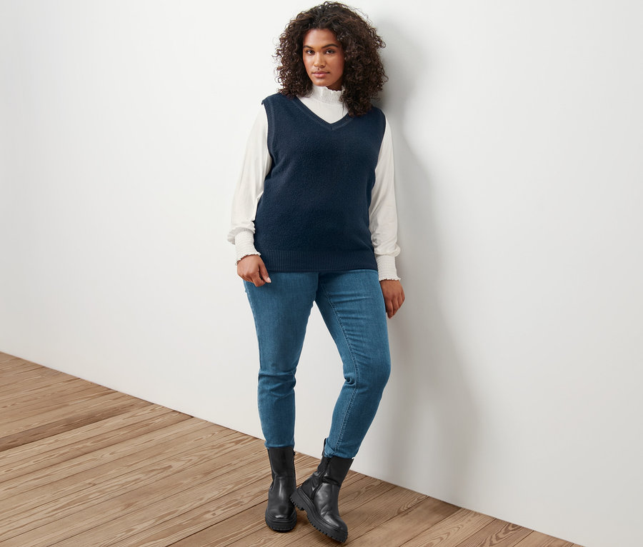 Žena pózuje v pletené vestě, krémovém halenovém triku se stojáčkem a tregínách »Fit Nelly«.