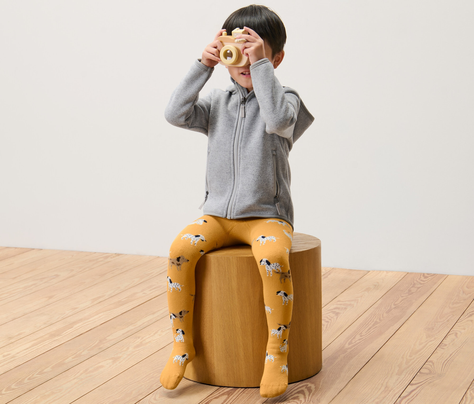 Chlapec sedí na stoličce a drží dřevěný fotoaparát před obličejem. Má na sobě zelené a žluté dětské termo punčocháče.