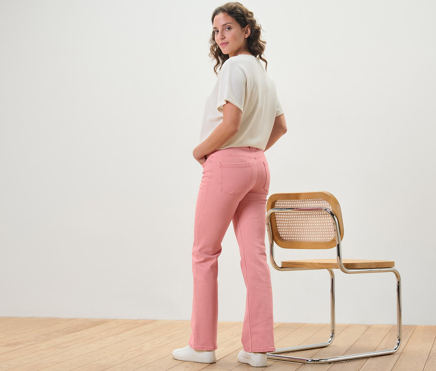 Žena pózuje v korálových džínách bootcut střihu »Lou« vedle židle.