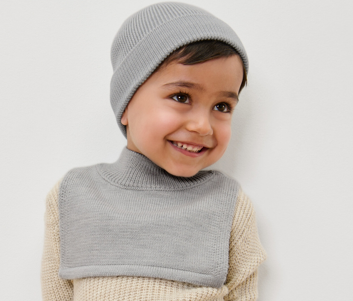 Usmívající se chlapec má na sobě šedou vlněnou rolákovou šálu a čepici.