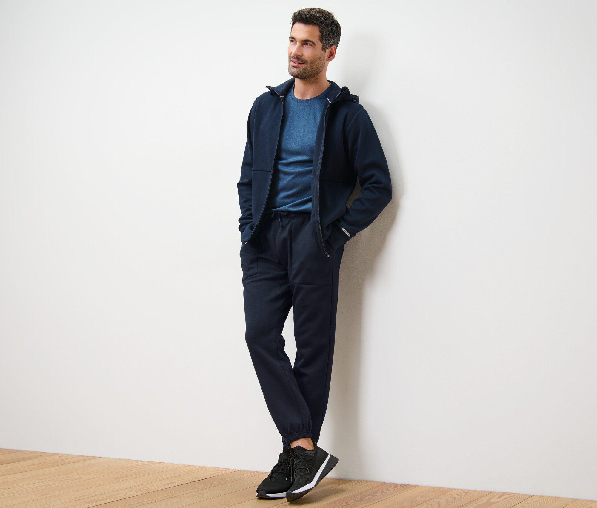 Muž opřený o zeď má na sobě tmavě modrou mikinu s kapucí, tričko a funkční teplákové kalhoty.
