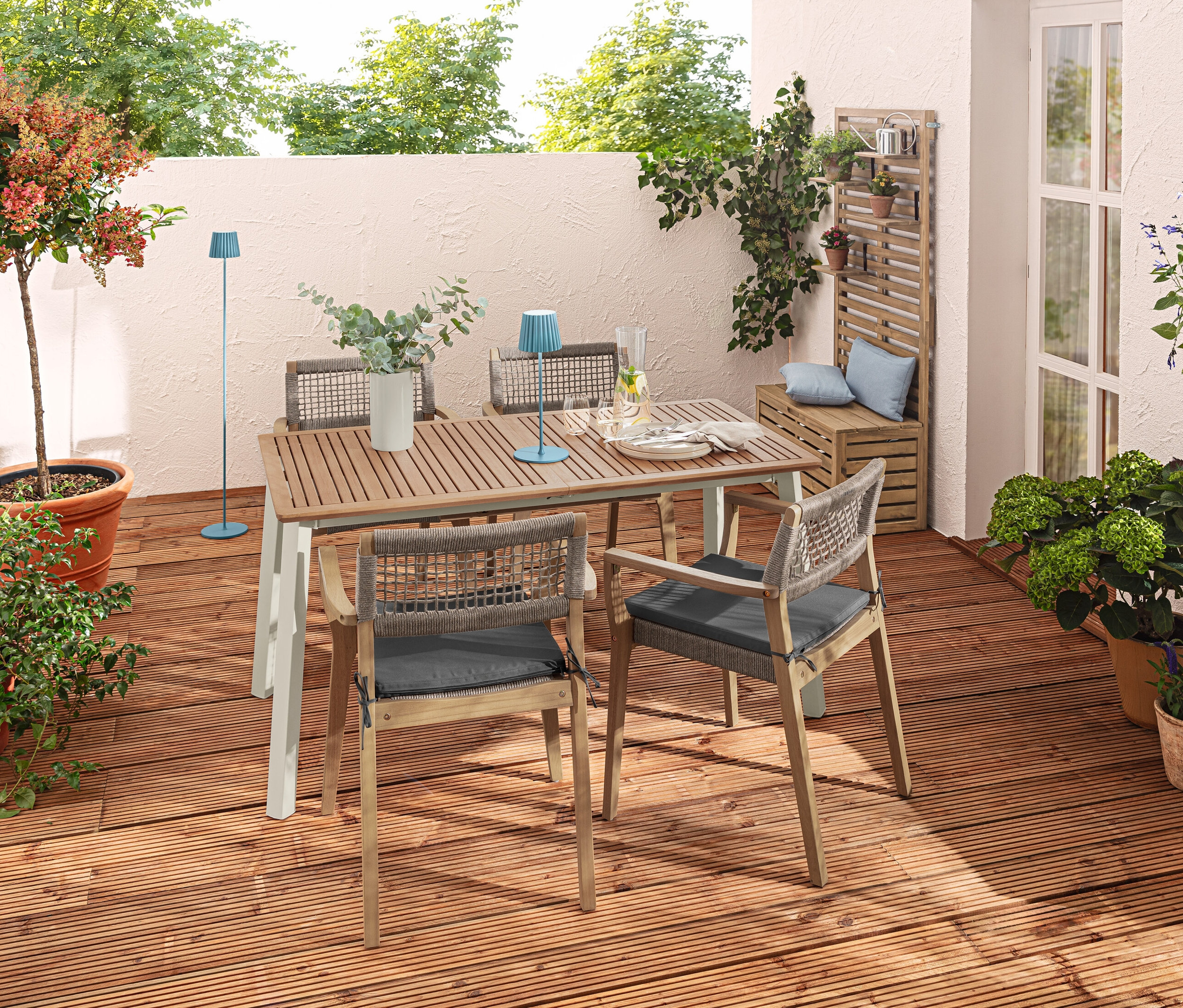 Venkovní terasa s rozkládacím stolem »Liska«, stohovatelnými židlemi, modrou LED stolní a stojací lampou a polštáři.