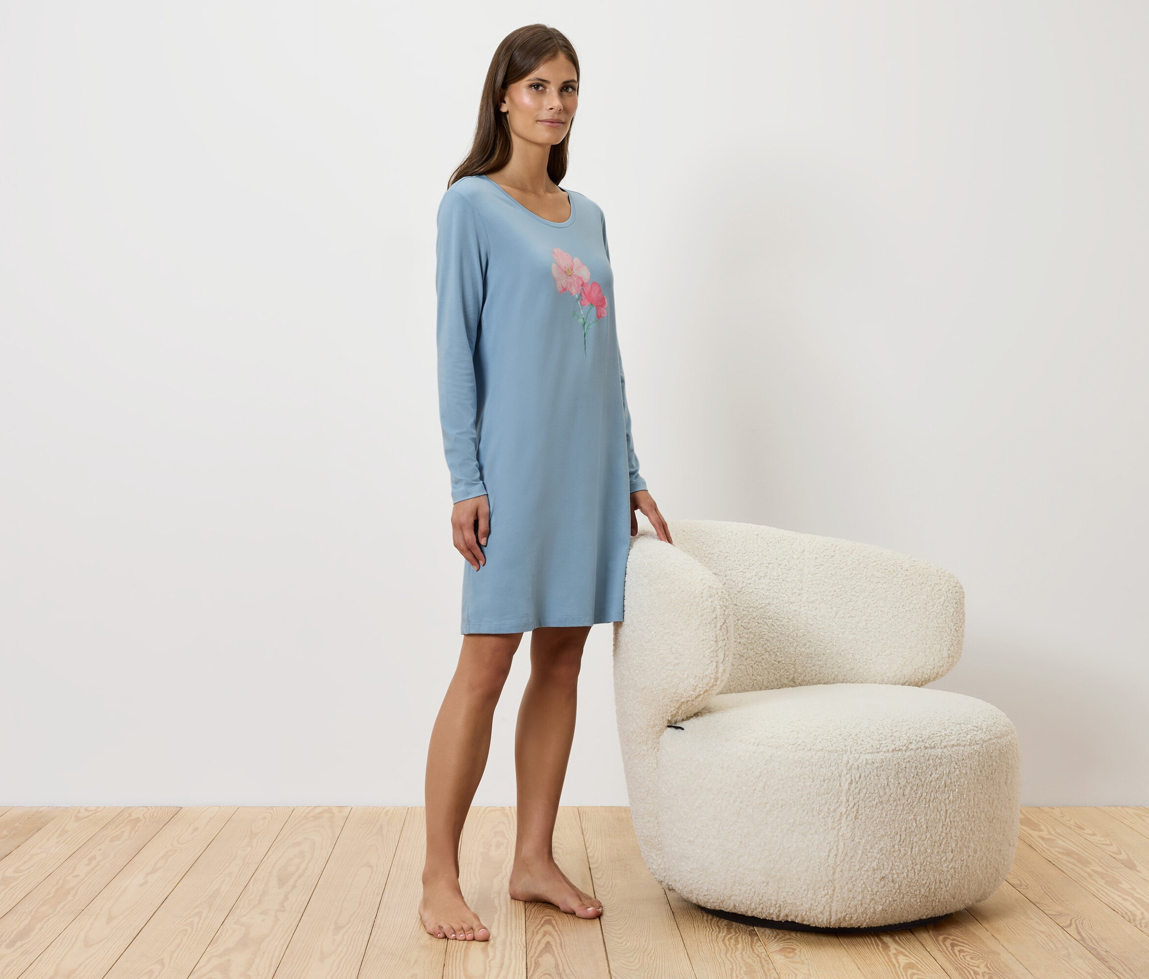 Žena stojí vedle bílého křesla a má na sobě modré noční šaty.