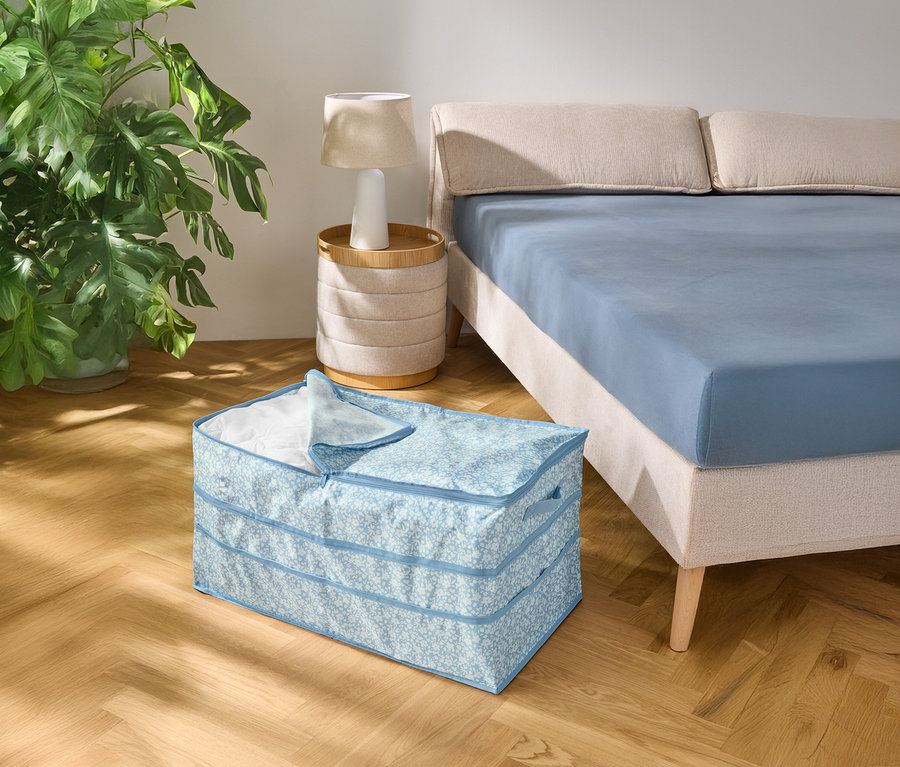 Na dřevěné podlaze stojí světle modrý vzorovaný úložný box vedle postele s modrým prostěradlem.