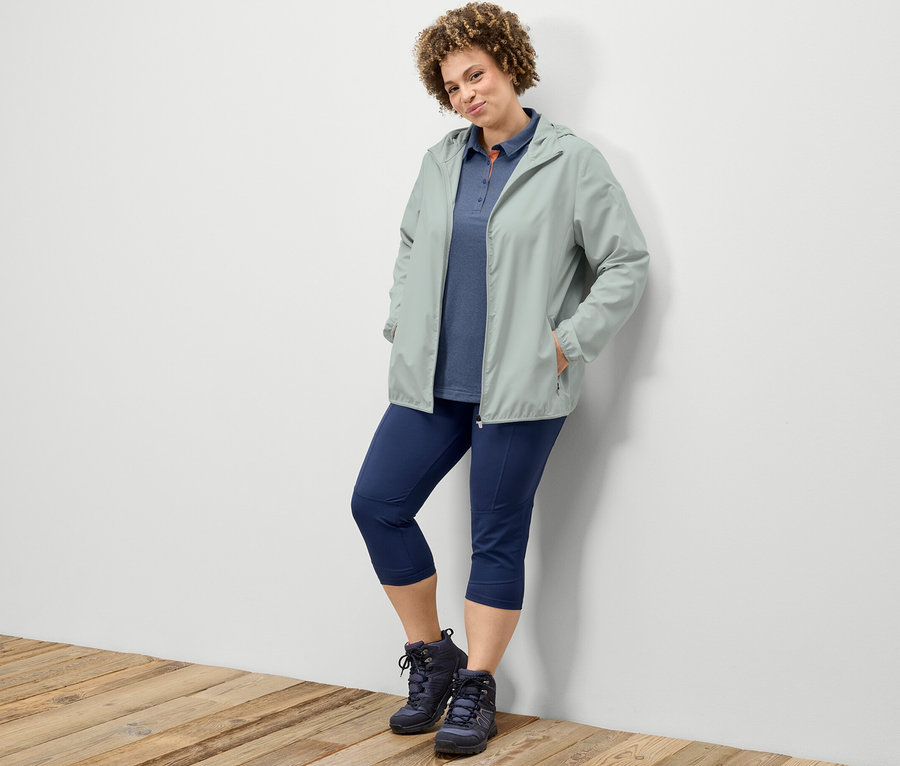 Žena opřená o zeď má na sobě 3/4 trekingové legíny, tmavě modrou funkční polokošili a šedou sbalitelnou funkční bundu.
