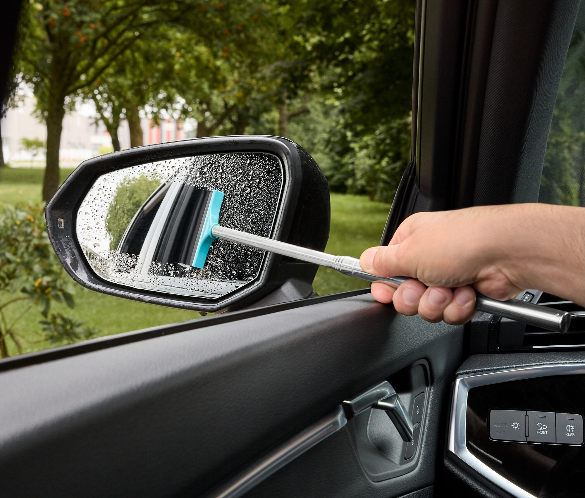 Muž stírá vodu z bočního zrcátka auta stěrkou s modrou gumou.