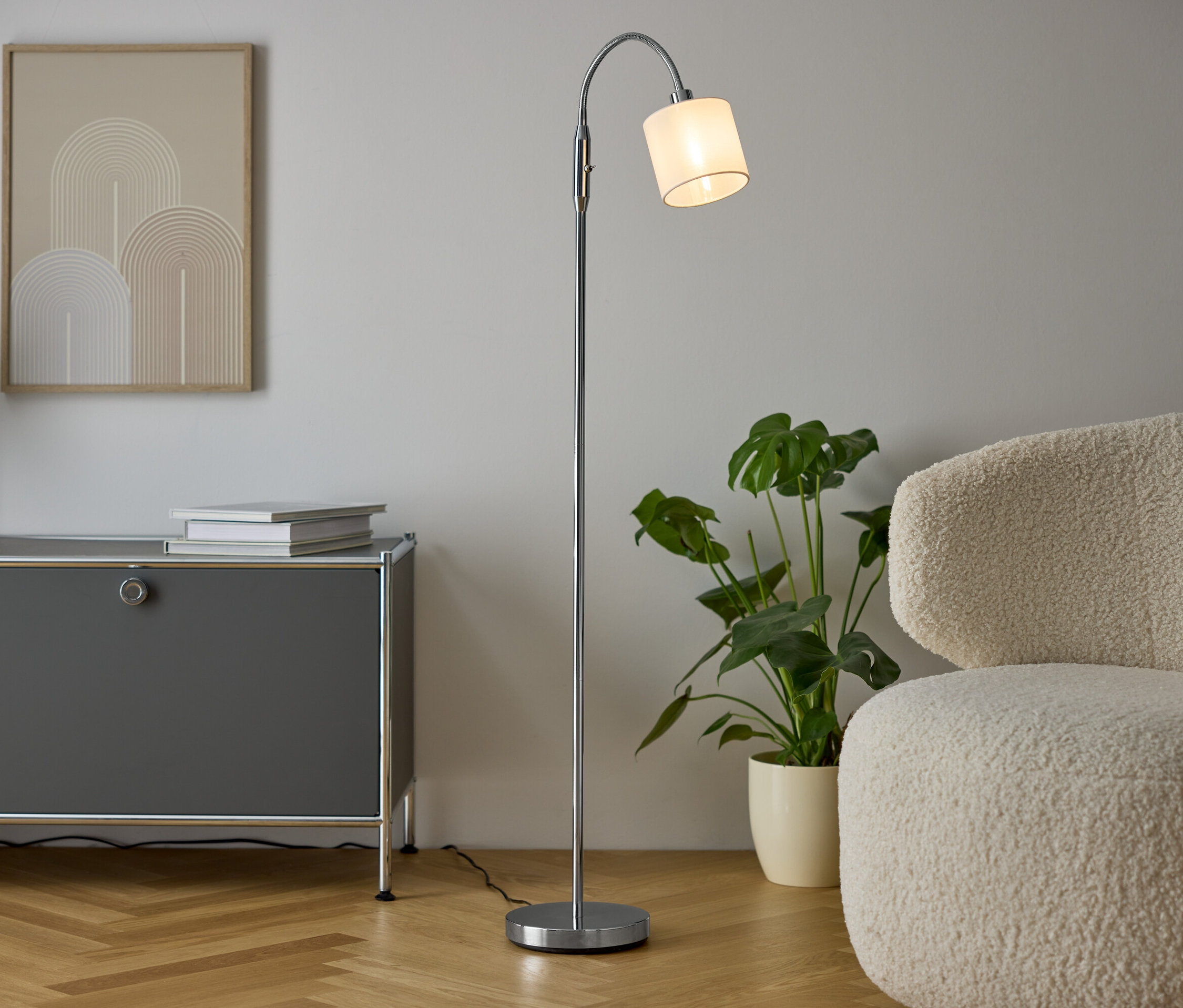 Interiér s šedou komodou, vázou se suchými květinami, dvěma knihami, stříbrnou lampou a krémovým křeslem.