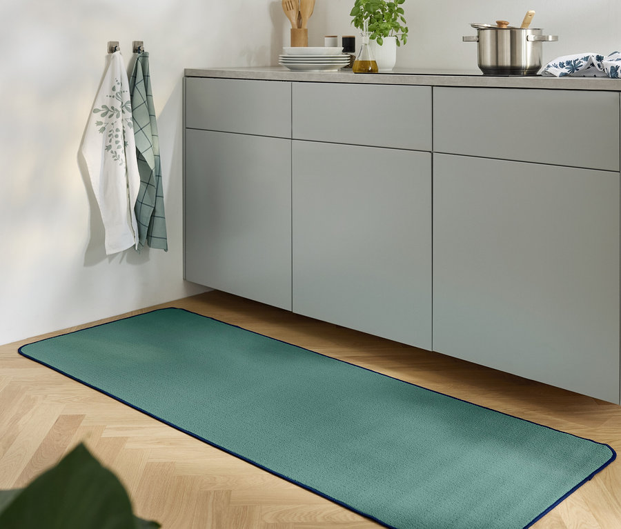 Zelený koberec leží na dřevěné podlaze v kuchyni s šedou skříňkou.