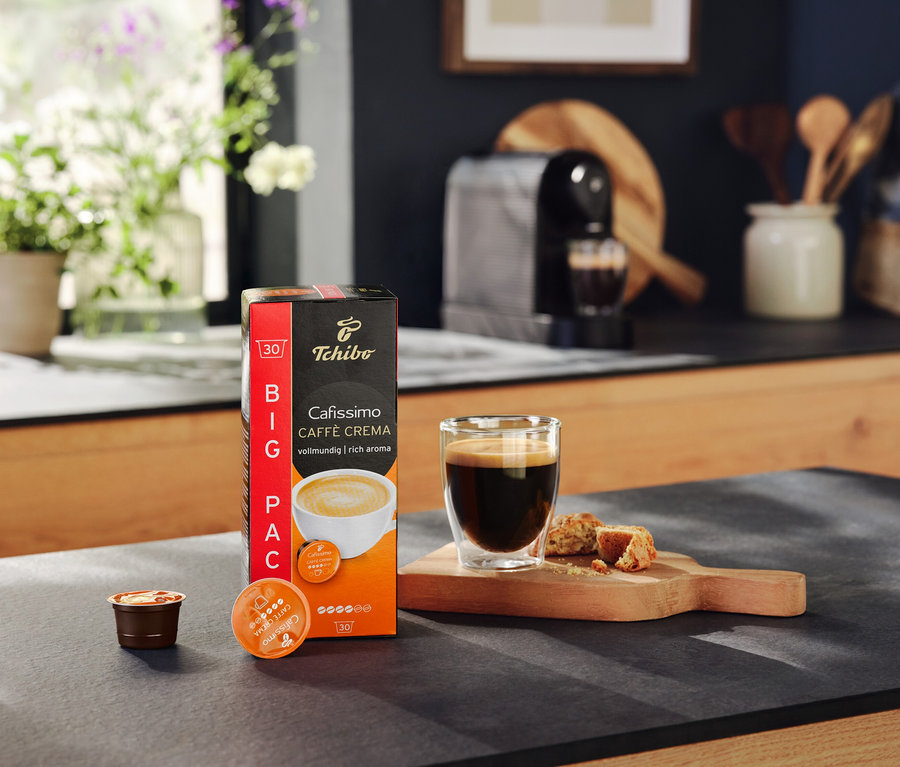 Balení kapslí Cafissimo Caffe Crema, sklenice s kávou a kousky sušenek na desce stolu. V pozadí kávovar Cafissimo pure+ black.