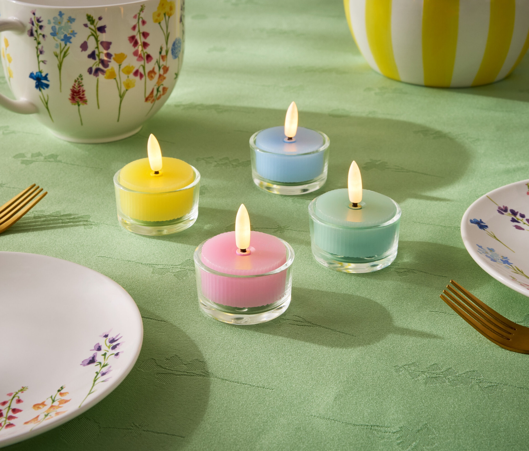 Na zeleném ubrusu jsou čtyři malé svíčky v pastelových barvách: žlutá, modrá, zelená a růžová. Dále je zde talíř, šálek a mísa s květinovým vzorem a dvě zlaté vidličky.