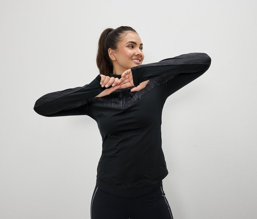 Usmívající se žena v černém sportovním oblečení se protahuje před bílým pozadím.