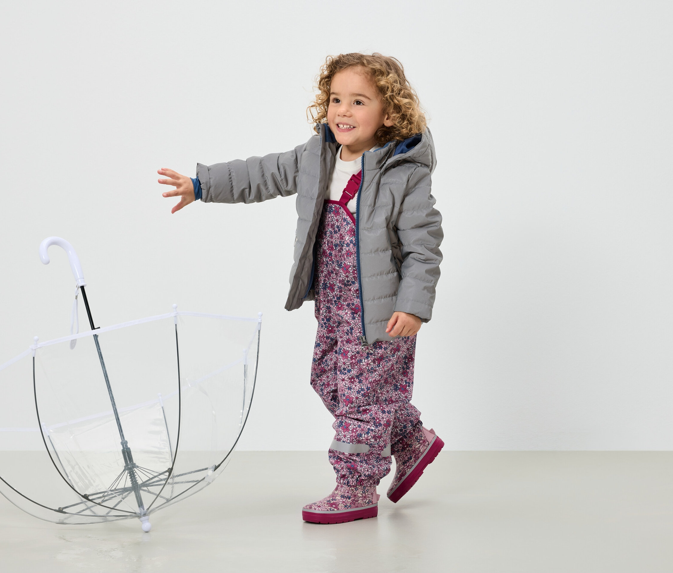 Usmívající se kudrnatá holčička v šedé bundě a květinových kalhotách stojí vedle průhledného deštníku.