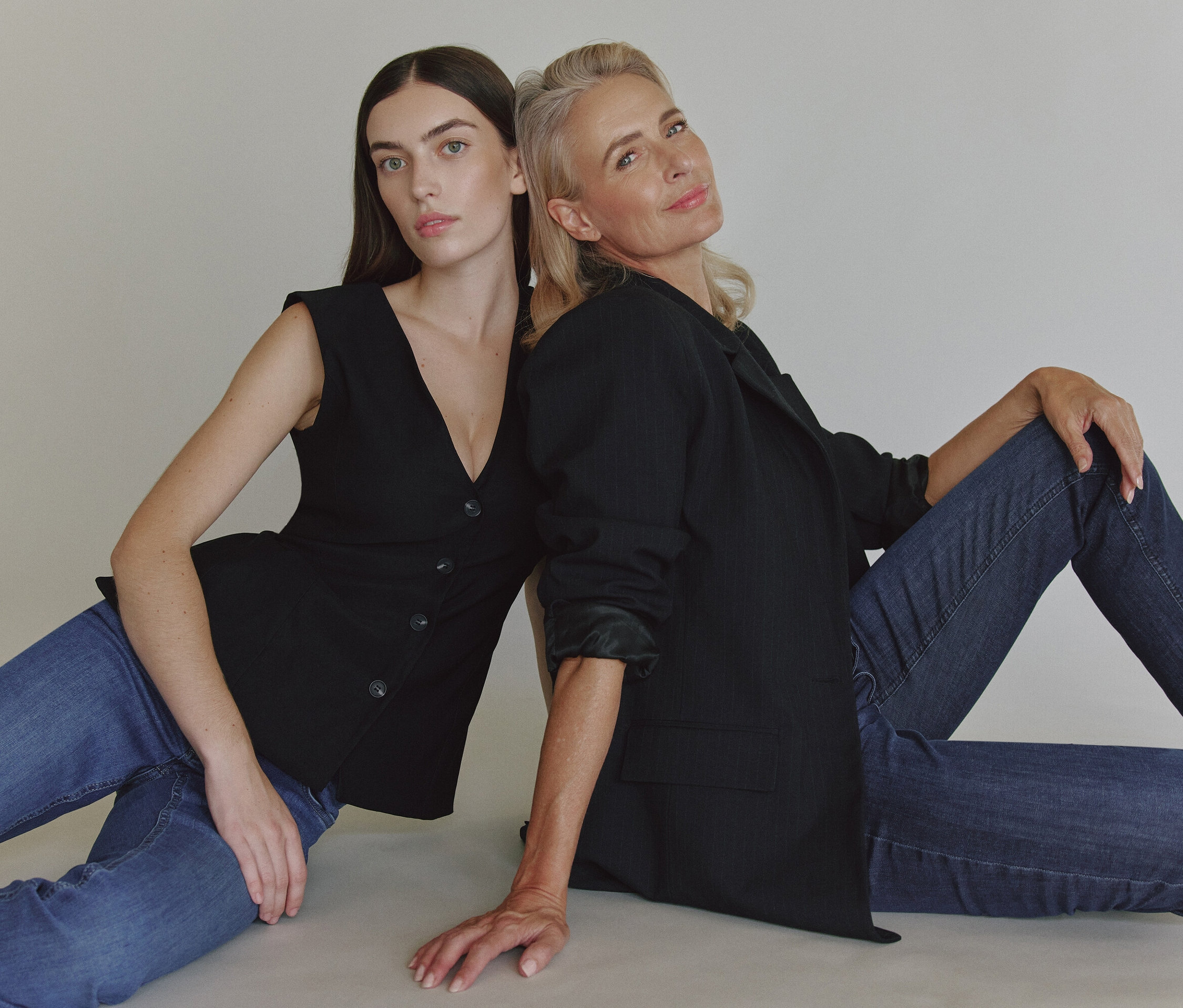 Dvě ženy sedí zády k sobě na podlaze. Jedna má na sobě černé sako a džíny, druhá černou vestu a džíny.