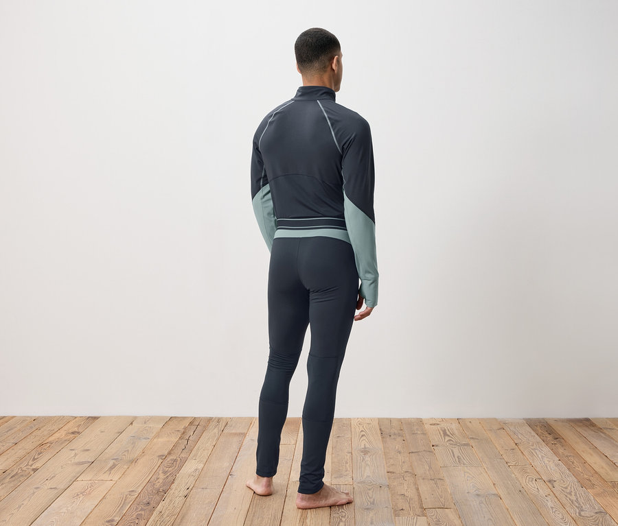 Muž zády ke kameře má na sobě tmavě šedé termoprádlo a stojí na dřevěné podlaze.