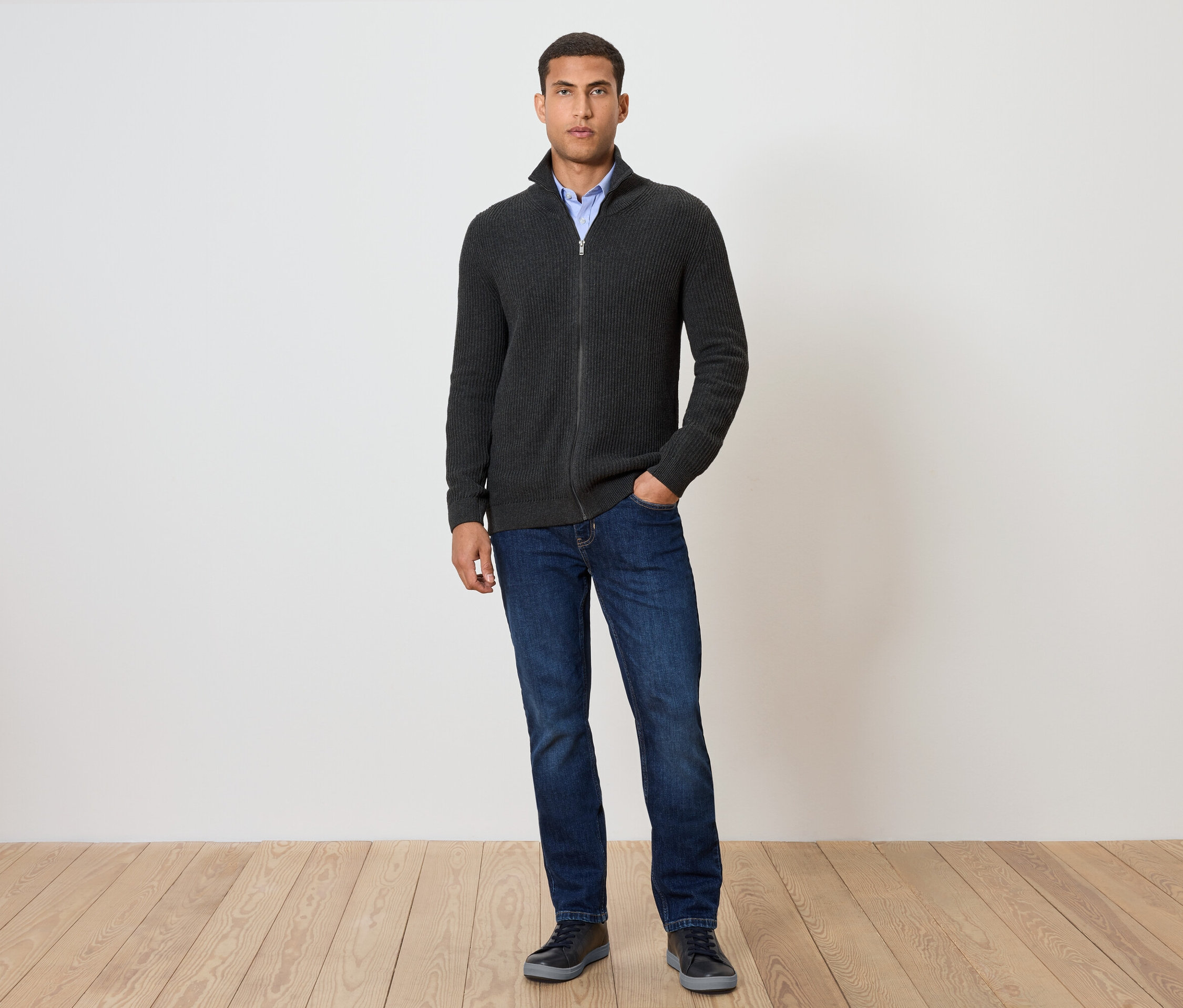 Muž stojí a má na sobě pletený svetr, antracitová s melírem, džíny a boty.