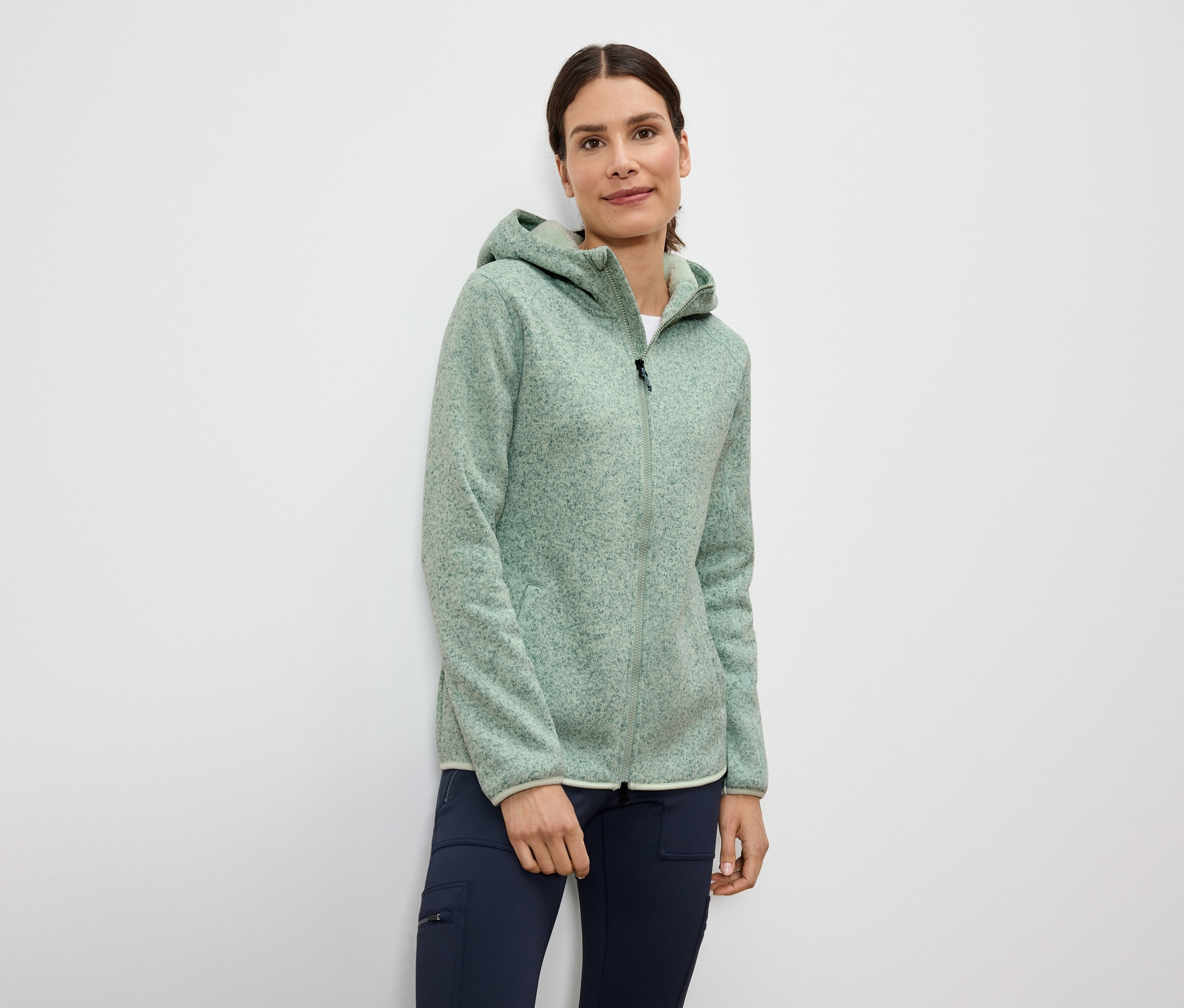 Žena má na sobě světle zelenou mikinu na zip s kapucí a tmavě modré kalhoty.