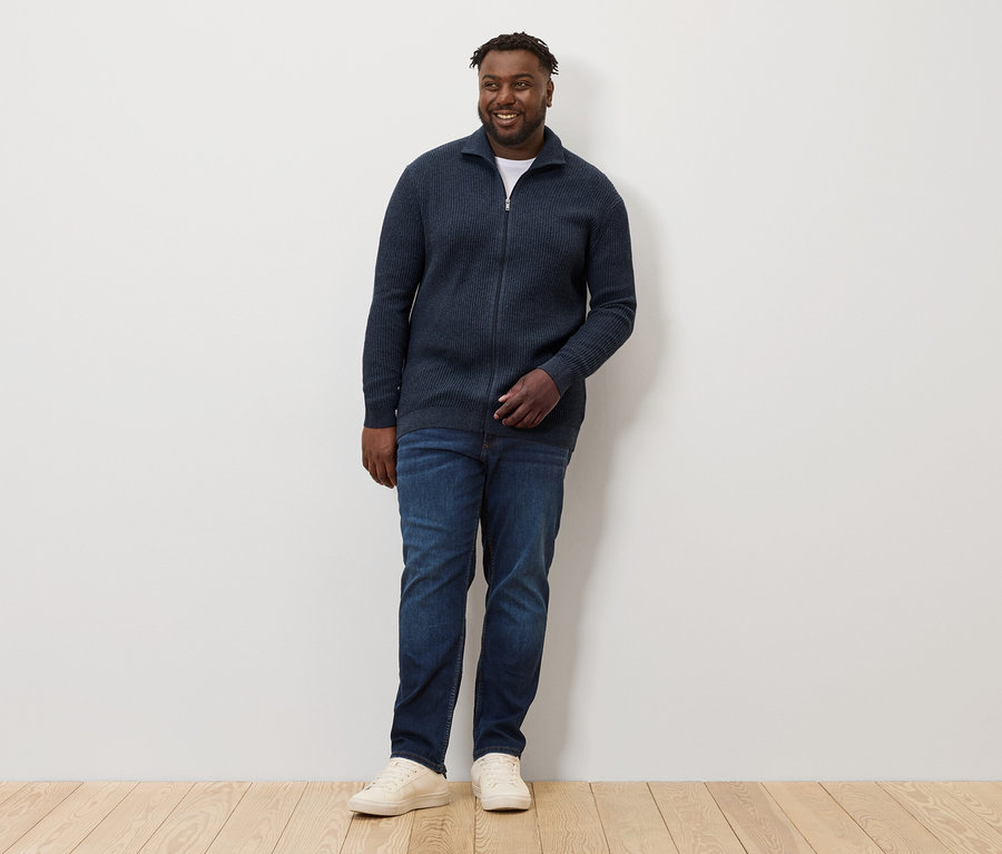 Muž stojí a má na sobě pletený svetr námořnické modré barvy s melírem, džíny a tenisky.