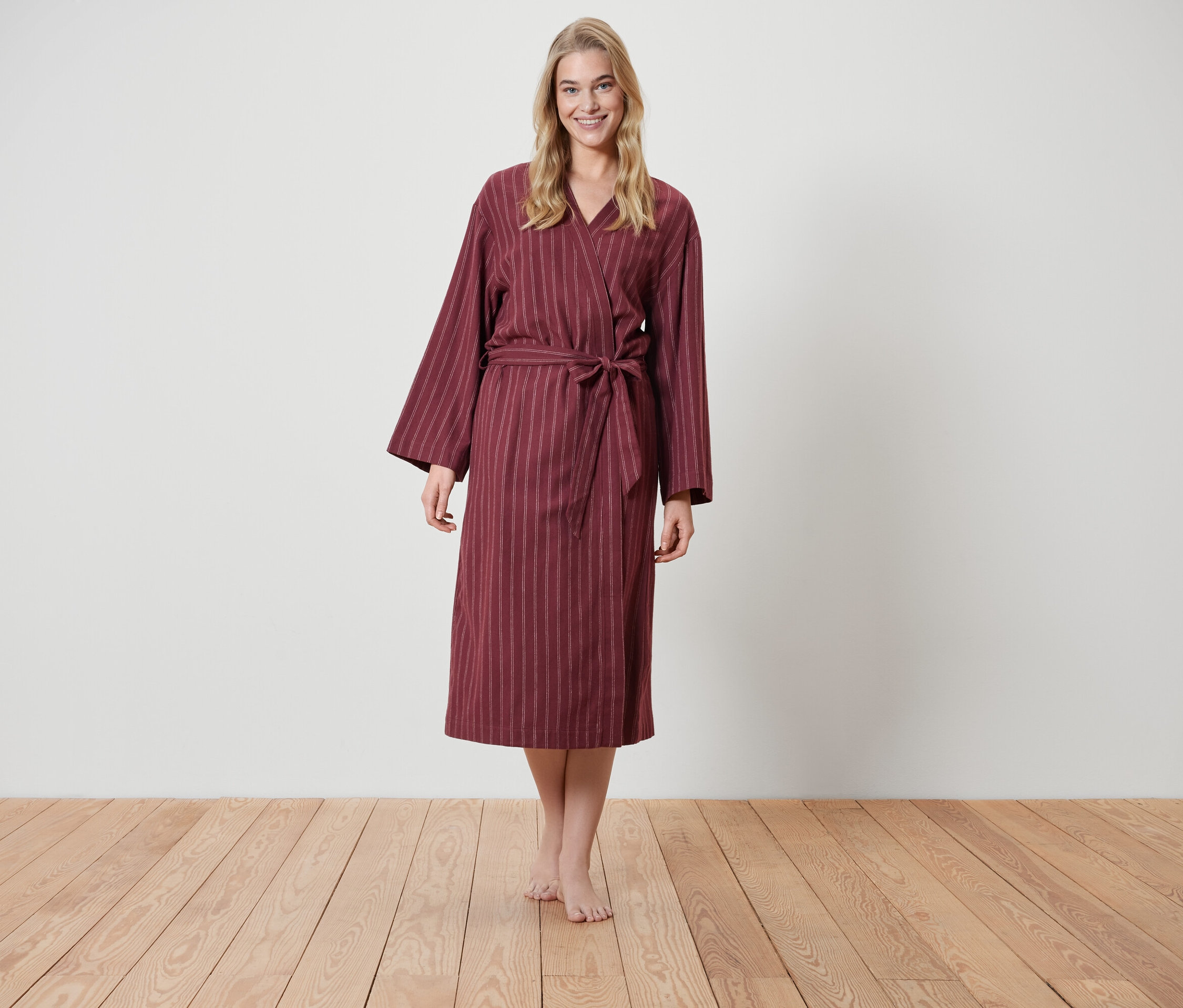 Blondýna stojí na dřevěné podlaze a má na sobě vínový župan s proužky.