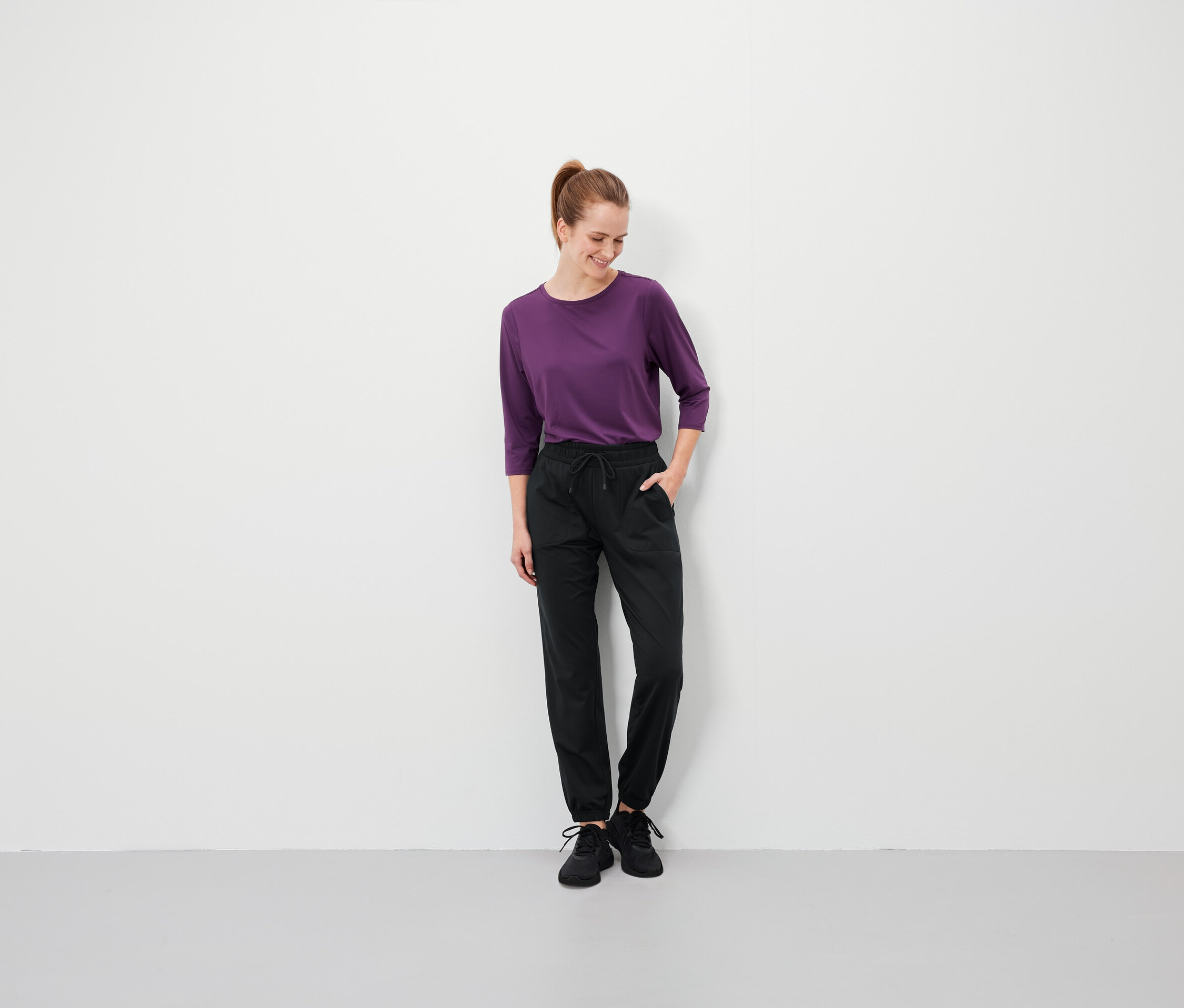 Žena s hnědými vlasy v culíku stojí a má ruce v kapsách černých kalhot a na sobě fialové tričko.