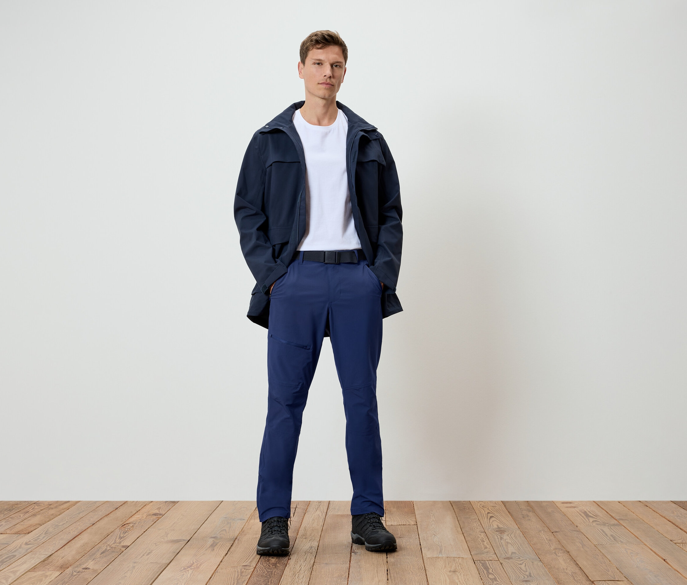 Muž stojí na dřevěné podlaze. Má na sobě tmavě modrou bundu, bílé tričko, modré kalhoty a černé boty.