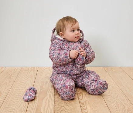 Kojenecká dívka sedí na dřevěné podlaze v květinovém zimním obleku s kapucí a jednou botou.