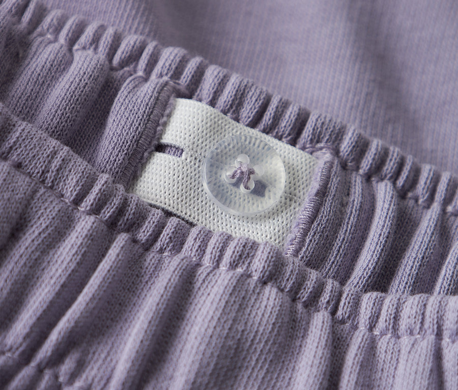 Detailní záběr na pas fialových kalhot s nastavitelnou gumou a knoflíkem.