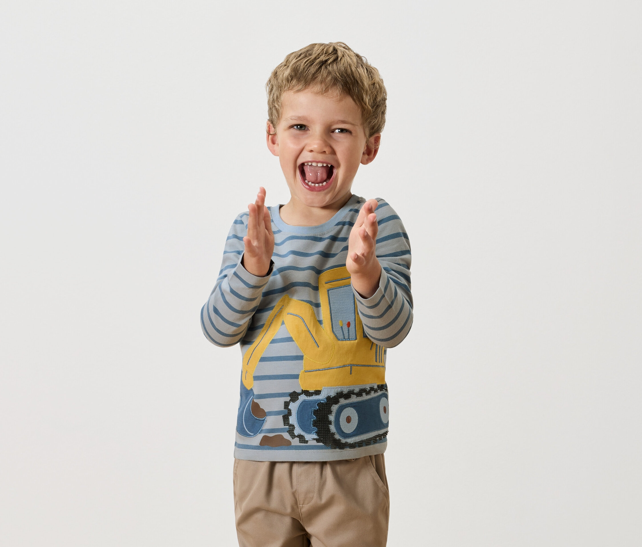 Chlapec s blond vlasy tleská rukama a má na sobě modře pruhované tričko s dlouhým rukávem se žlutým buldozerem.