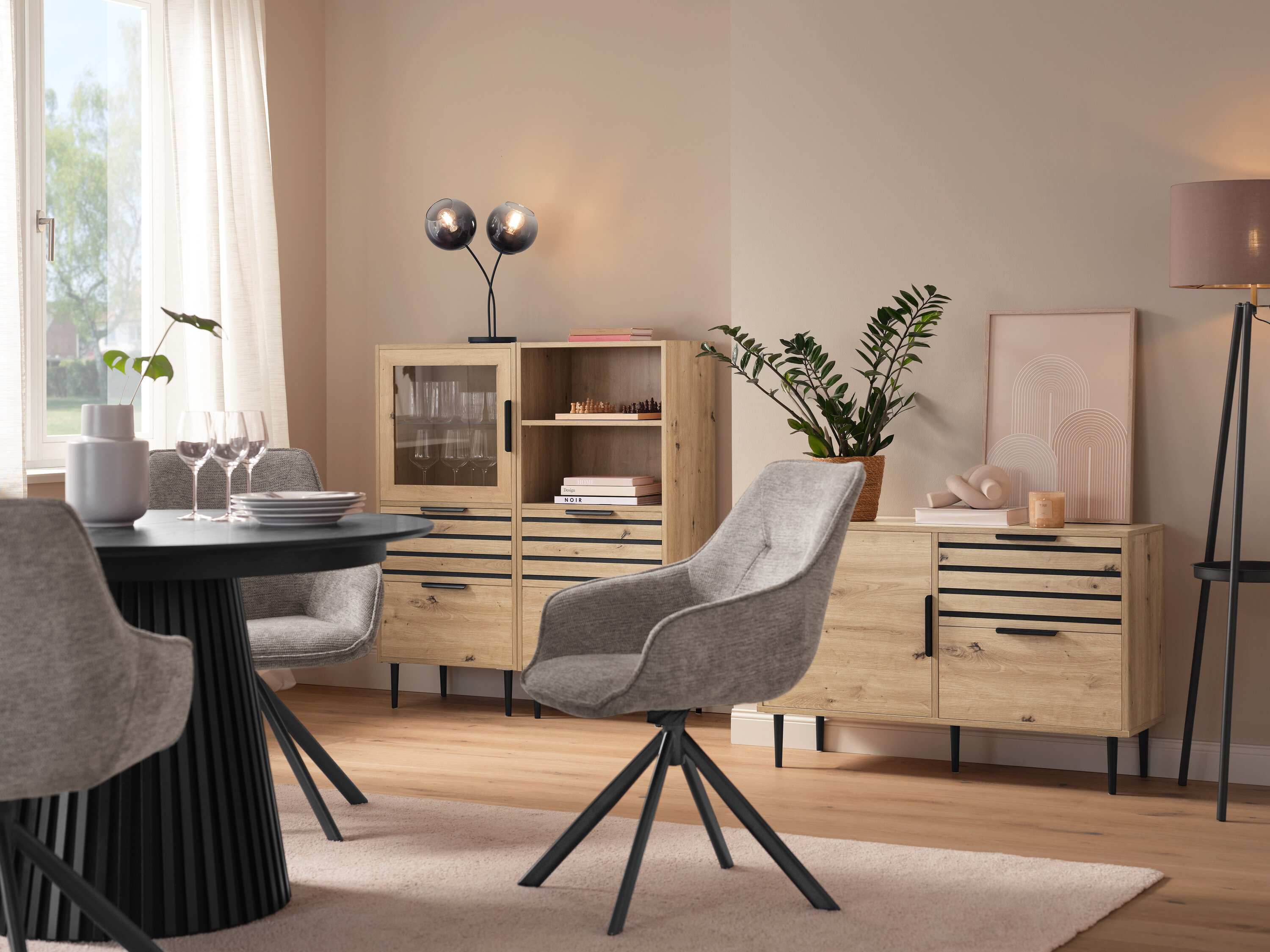 Jídelna s kulatým stolem, šedými židlemi, vitrínou »BAAKEN« se skleněnými dvířky, regálem »BAAKEN« se zásuvkami a sideboardem se zásuvkami.