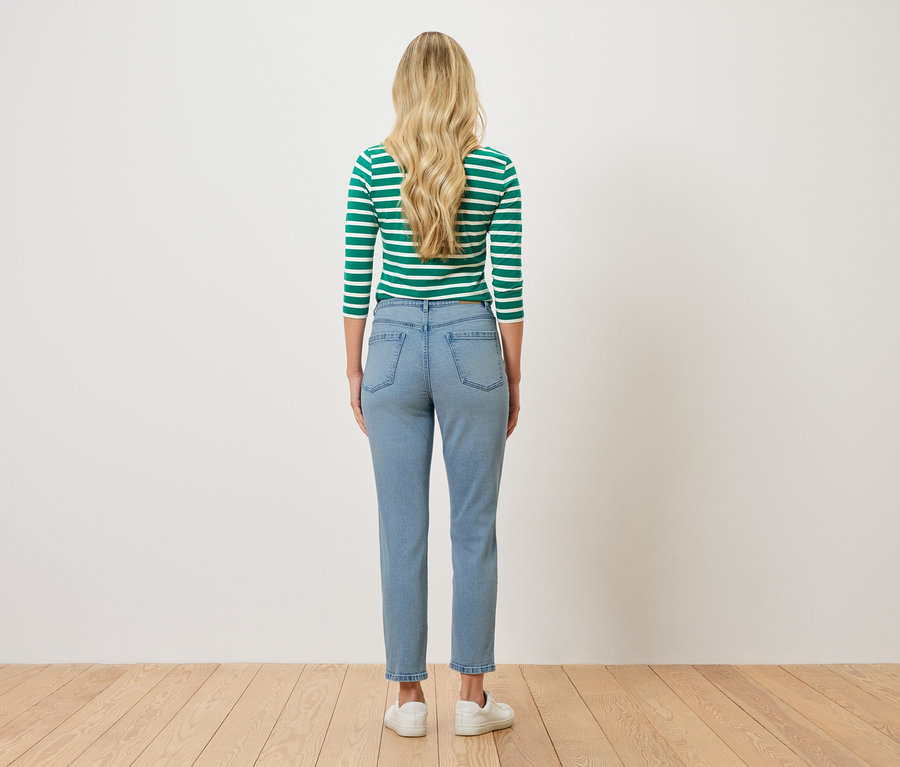 Pohled zezadu na ženu s blond vlnitými vlasy, oblečenou do zeleno-bílého pruhovaného topu, světle modrých džínů a bílých tenisek.
