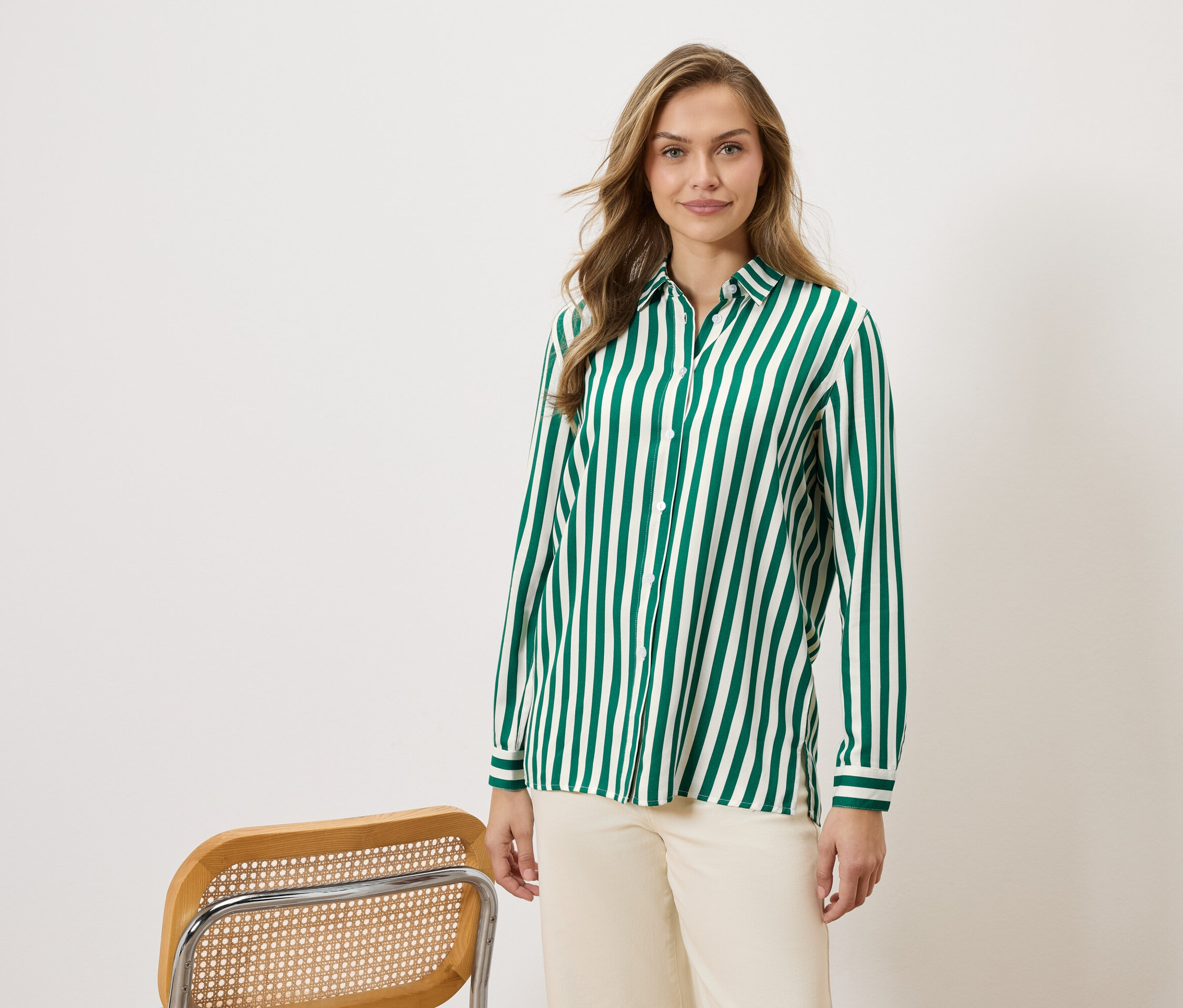 Žena má na sobě zeleno-bílou pruhovanou košili a béžové kalhoty, vedle židle.
