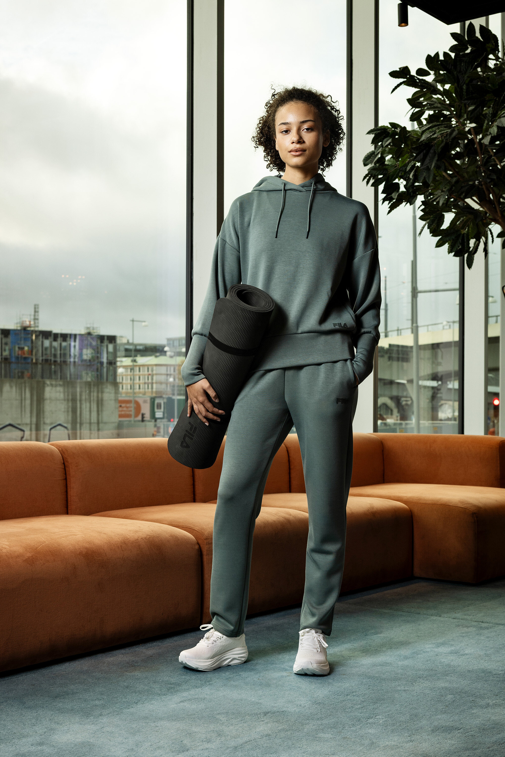 Mladá žena v šedém sportovním oblečení drží srolovanou podložku na jógu před oknem.