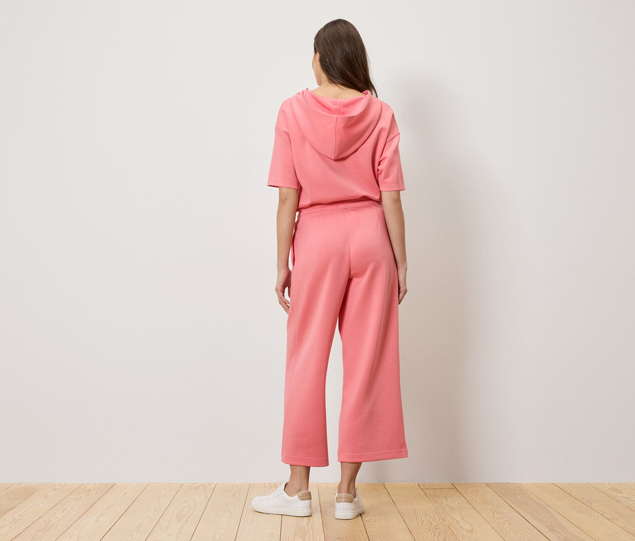 Žena stojí zády ke kameře a má na sobě růžovou mikinu s kapucí a culottes kalhoty.