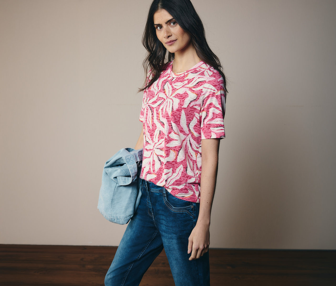 Žena má na sobě růžové tričko s krátkým rukávem CECIL se vzorem a džíny. Na rameni má džínovou bundu.