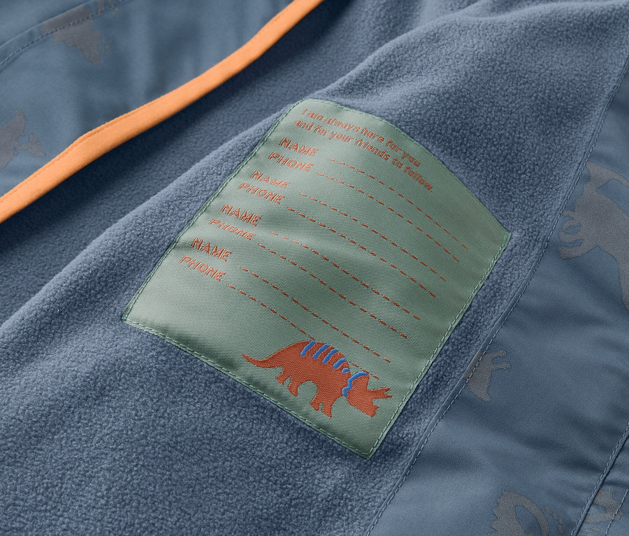 Detailní pohled na vnitřek modré bundy s oranžovým lemem a štítkem s dinosaurem a prostorem pro jméno a telefonní číslo.