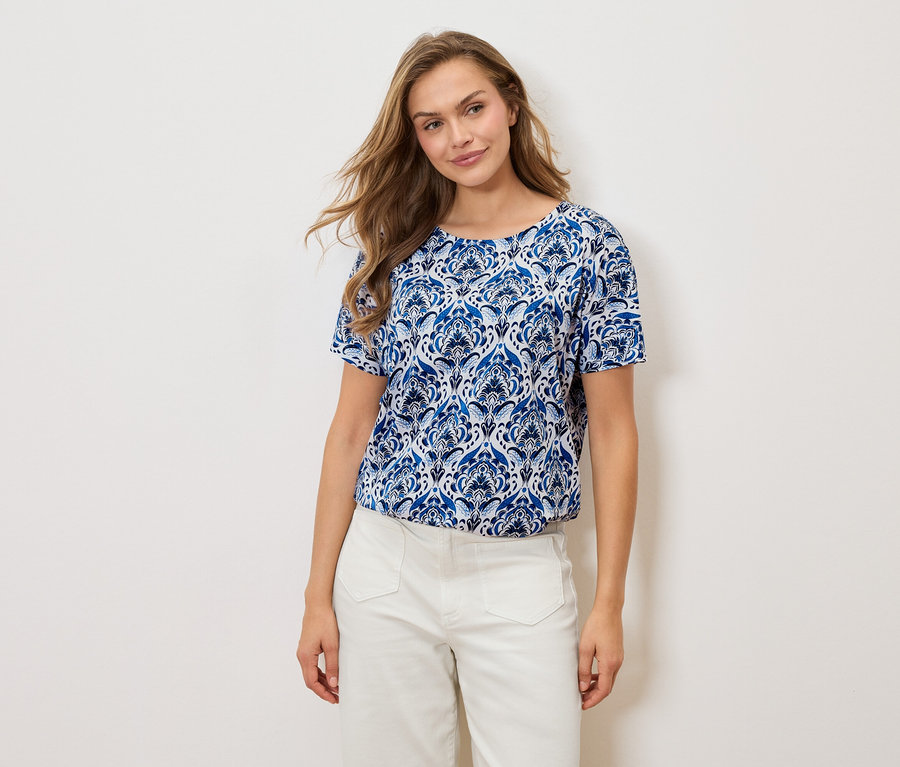 Žena má na sobě modrobílé vzorované tričko a bílé kalhoty na bílém pozadí.