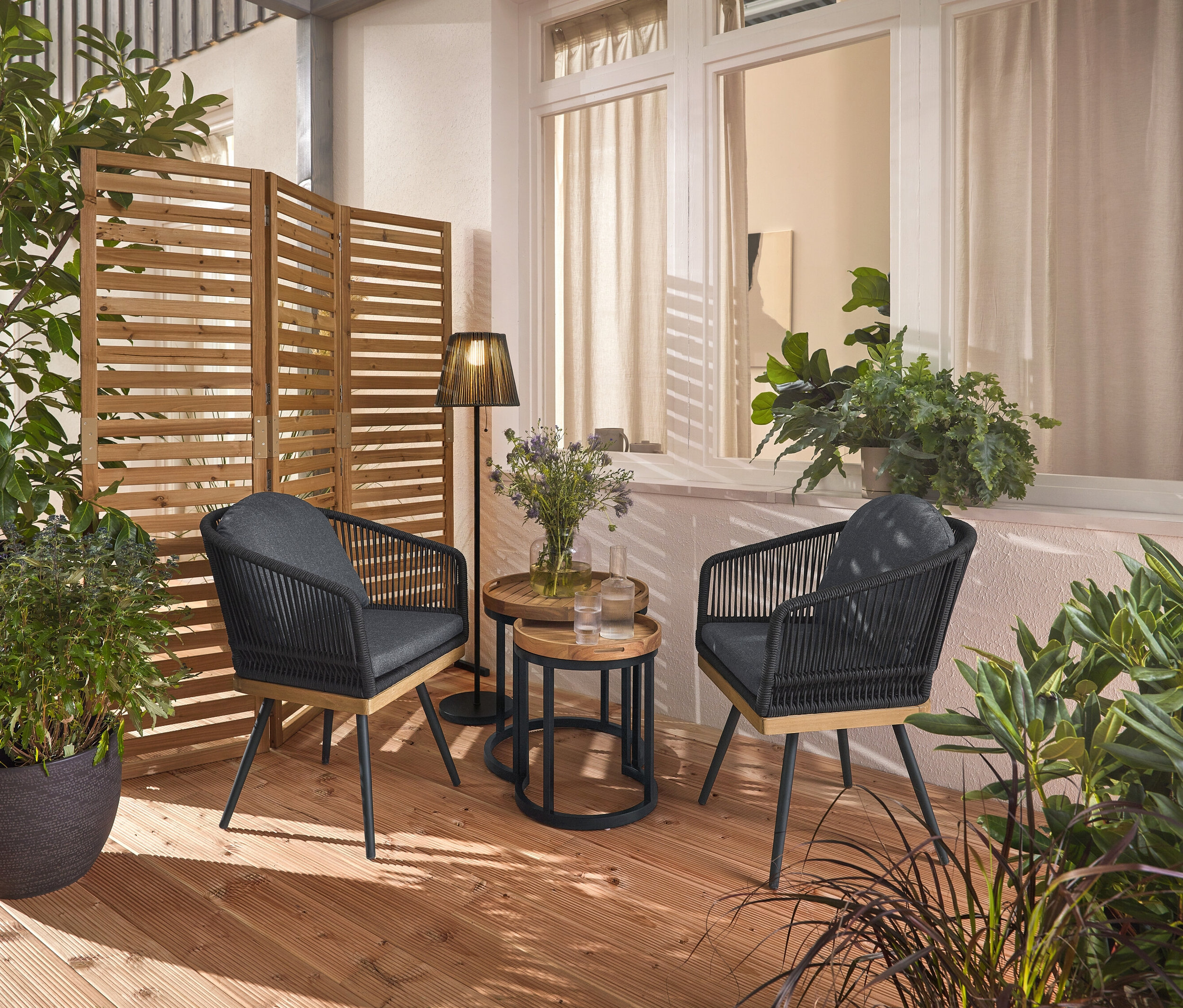 Dvě otočné zahradní židle s textilním výpletem stojí na dřevěné terase, mezi nimi dva zahradní odkládací stolky s odnímatelnými podnosy, průměr cca 40 cm a 50 cm. V pozadí flexibilní paraván a dobíjecí solární stojací lampa.