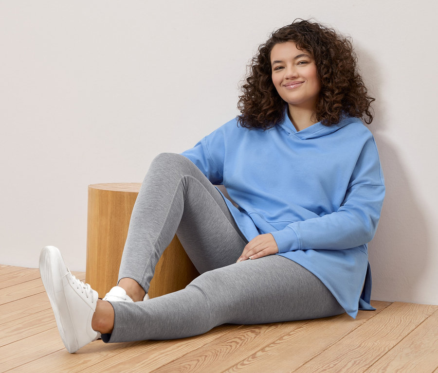 Žena s kudrnatými vlasy sedí na zemi, má na sobě světle modrou mikinu s kapucí, šedé legíny a bílé tenisky.