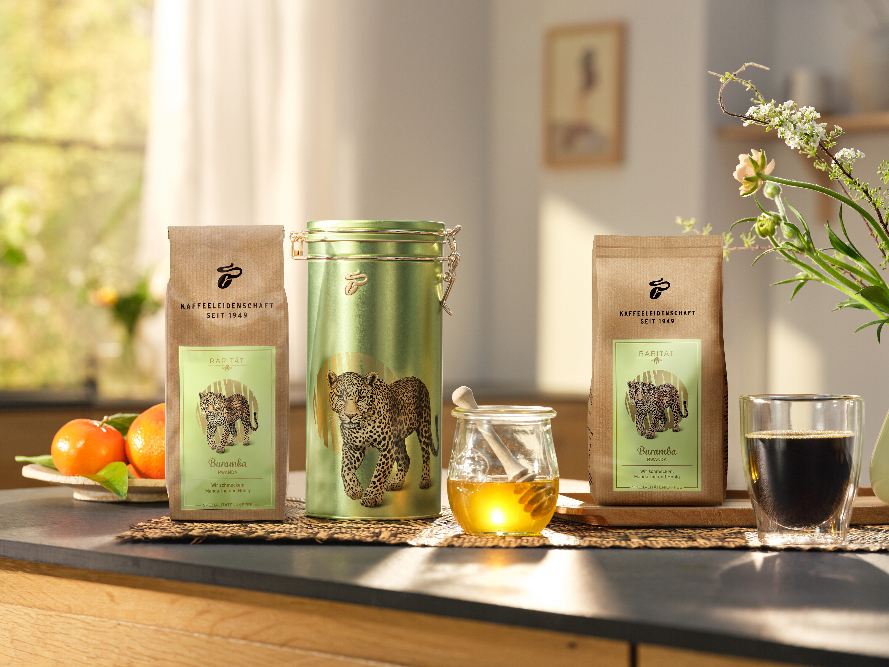 Na kuchyňské lince stojí dóza na kávu a dvě balení »Buramba Rwanda« včetně dózy na kávu vedle šálku kávy a medu.