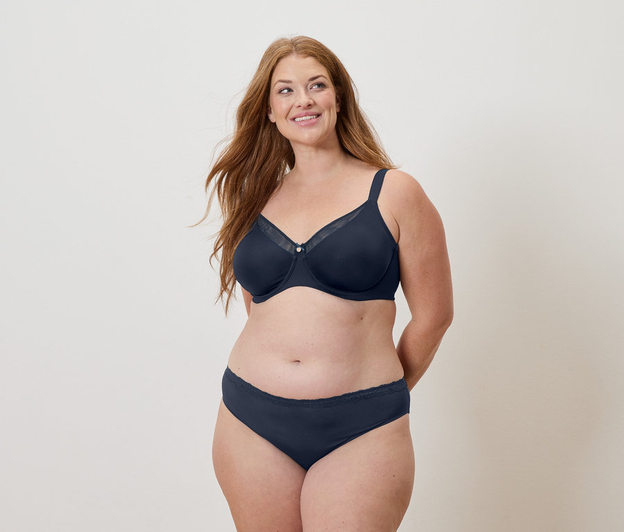 Žena s dlouhými zrzavými vlasy má na sobě tmavě modrou podprsenku a kalhotky a usmívá se.