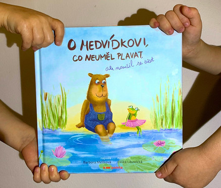 Dětské ruce drží knihu „O medvídkovi, co neuměl plavat, ale naučil se číst“ s obalem s medvědem a žábou.