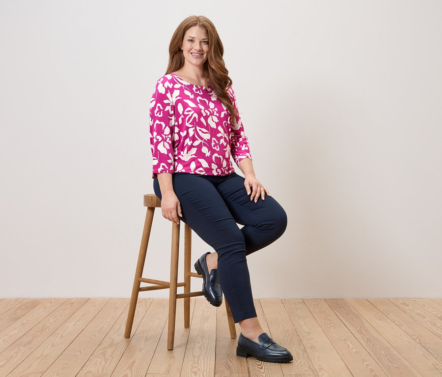 Žena sedí na stoličce, má na sobě růžovou blůzu s květinovým vzorem, modré kalhoty a kožené boty.