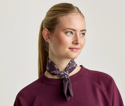 Žena s blond vlasy má na sobě vínovou mikinu a vzorovaný šátek.