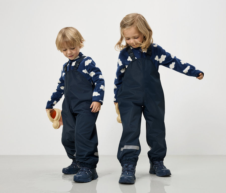 Dvě děti v tmavě modrých kombinézách a modro-bílých svetrech s mráčky stojí na bílém pozadí.