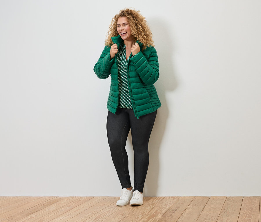 Usmívající se žena s kudrnatými vlasy v zelené prošívané bundě, pruhované halence, tmavých legínách a bílých teniskách.