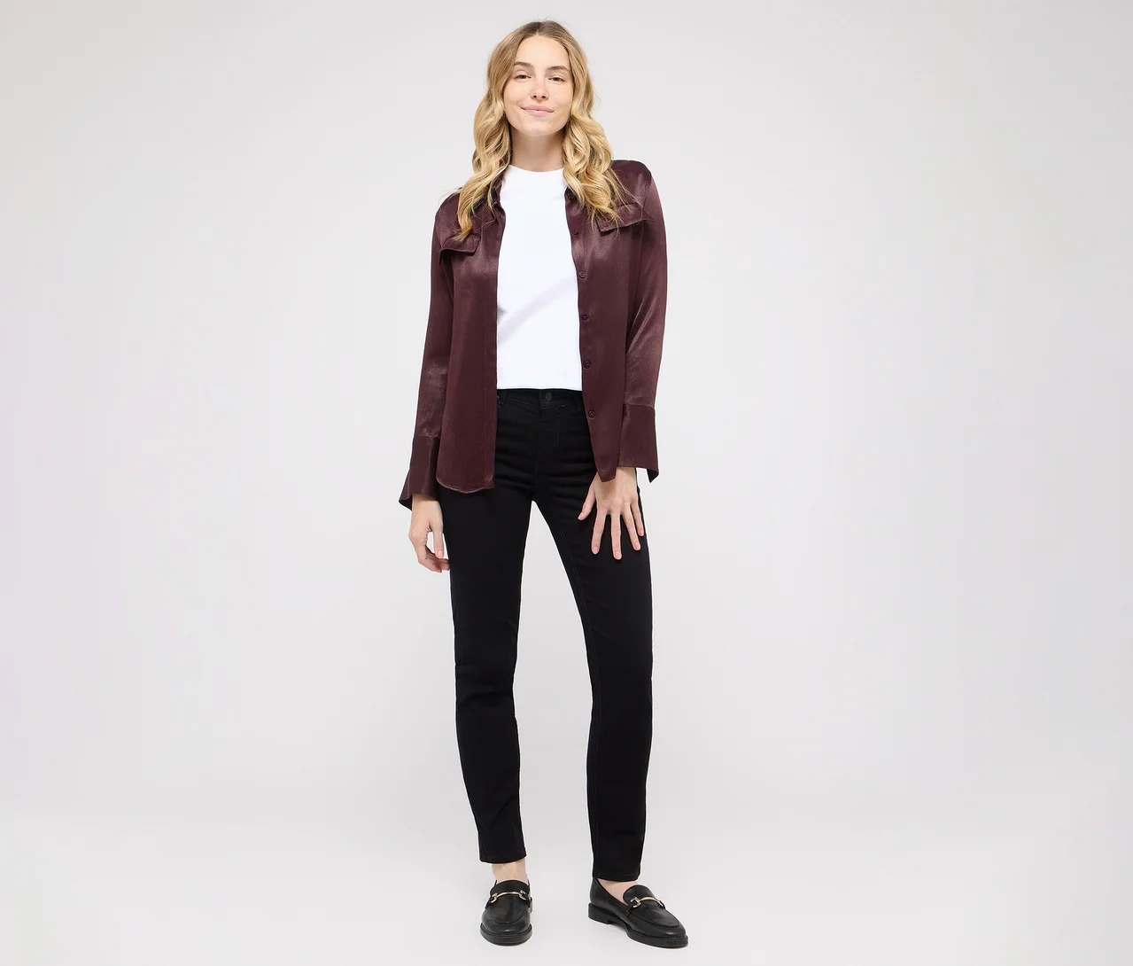 Žena s blond vlasy stojí a usmívá se před bílým pozadím. Má na sobě bílé tričko, otevřenou bordó košili, černé skinny džíny a černé mokasíny se zlatými sponami.