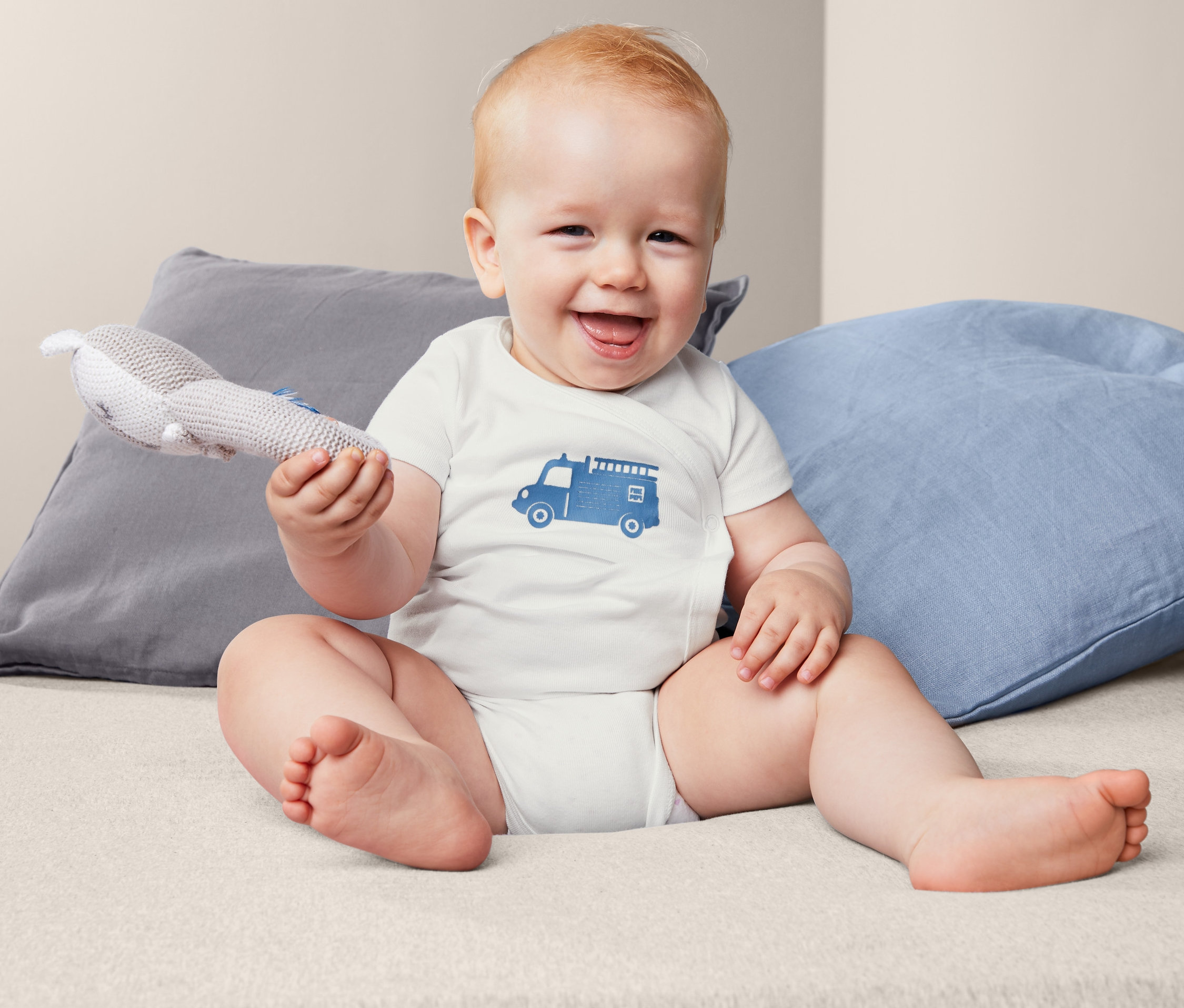 Usmívající se dítě sedí na posteli a má na sobě body s krátkým rukávem s potiskem hasičského auta a drží hračku.