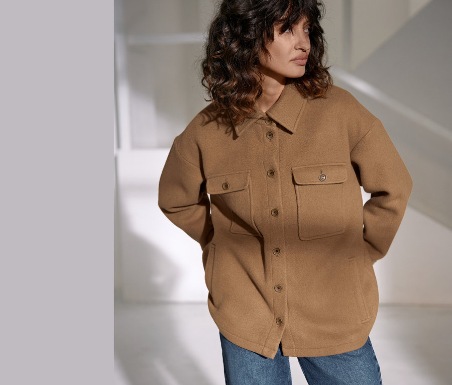 Žena má na sobě bundu NAH/STUDIO z recyklované vlny velbloudí barvy a džíny.