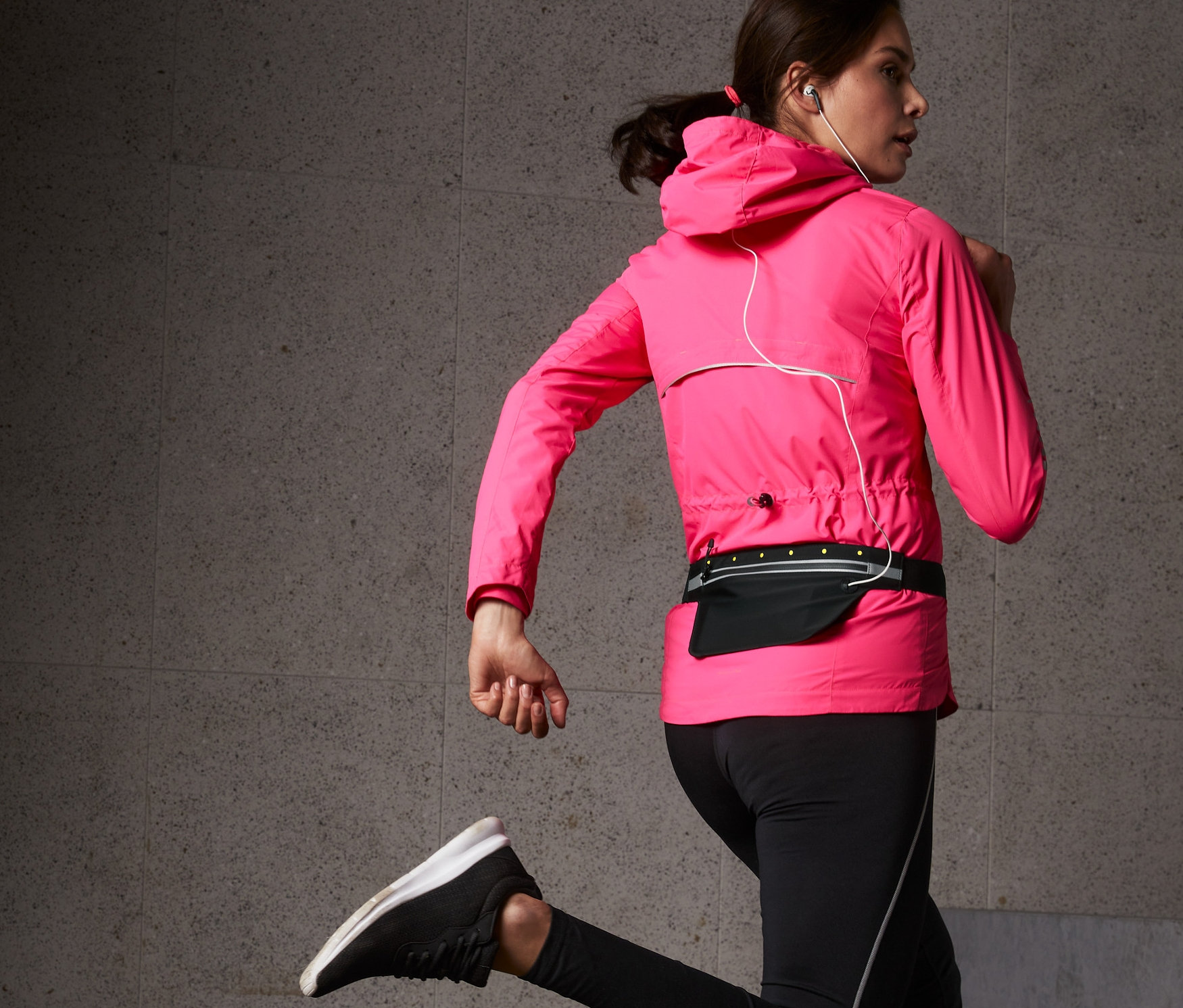 Žena v růžové bundě a černých legínách běží s černým běžeckým pásem s LED na smartphone.
