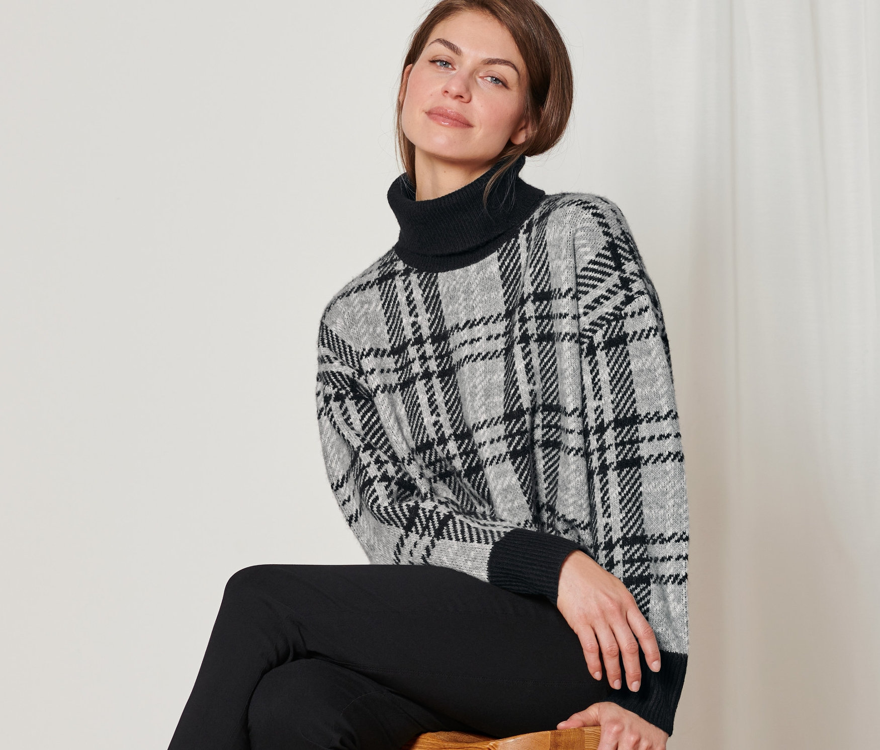 Žena sedí na stoličce a má na sobě pletený svetr s rolákovým límcem.